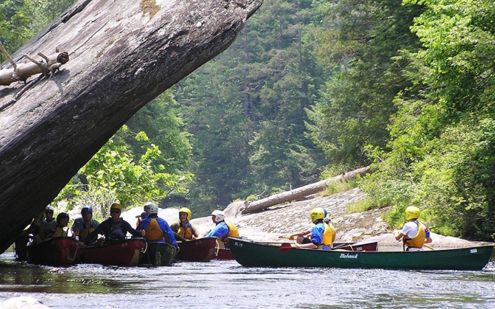 students sitting in canoes pause for a break in the shade of a large rock